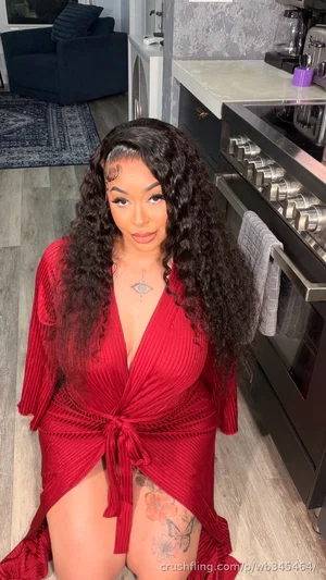 Woman with long curly hair in red wrap dress, standing in kitchen, showing thigh tattoo.