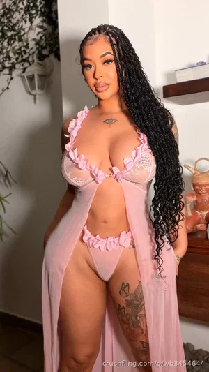 Woman with long black braids in pink lace lingerie and sheer robe, standing indoors.