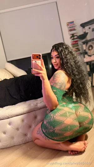 Woman with long curly hair in green sheer dress, kneeling on floor in bedroom, taking mirror selfie.