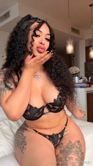 Woman with curly black hair and tattoos wearing black lace lingerie, posing on a bed indoors.