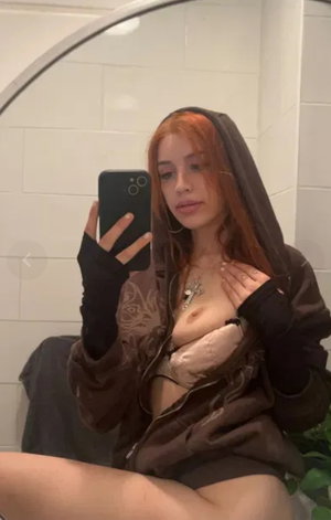 Red-haired woman in open hoodie taking mirror selfie in bathroom.