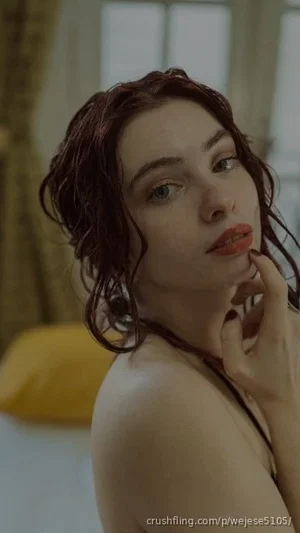 Woman with wet dark hair, bare shoulders, posing indoors near a bed.