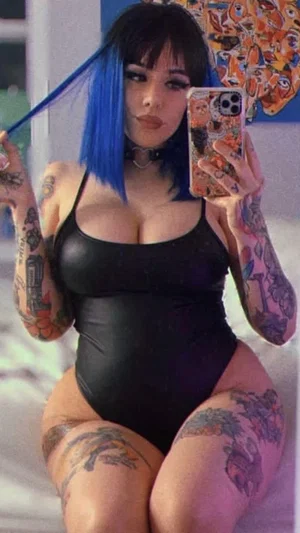 Woman with blue hair and tattoos wears black bodysuit, takes mirror selfie in bedroom.