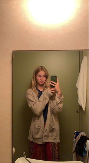 Woman with long blonde hair in beige cardigan taking bathroom mirror selfie.