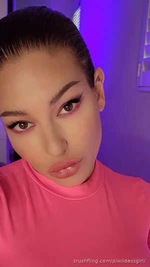Woman with dark hair pulled back, wearing a pink high-neck top, in a room with purple lighting.