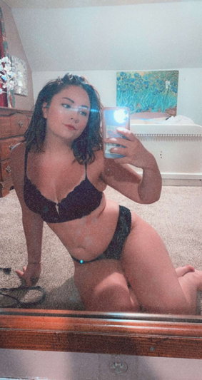 Woman in black lingerie posing on carpet in bedroom mirror selfie.