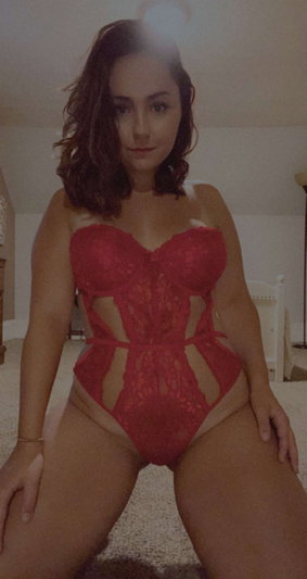 Woman in red lace bodysuit posing on carpeted floor indoors.