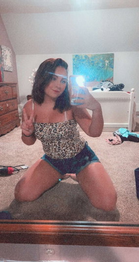 Woman in leopard top and shorts kneeling on carpet in bedroom, taking mirror selfie.