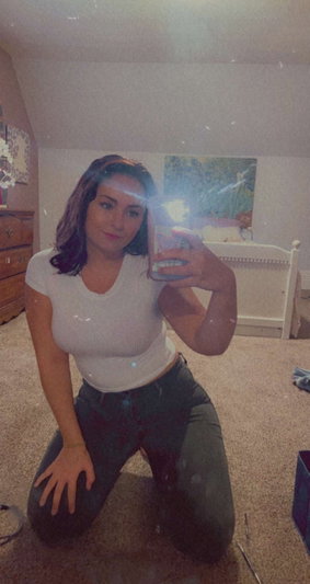 Woman in white top and dark pants kneeling in bedroom, taking mirror selfie.