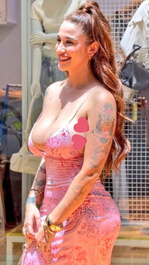 Woman with tattoos in pink dress, smiling, standing outdoors near store window.
