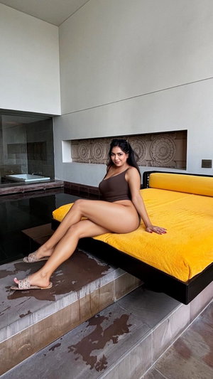 Woman in brown bodysuit sitting on yellow bed in modern indoor space.