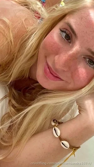 Blonde woman with freckles wearing a white top and shell bracelet, resting her head on her hand.