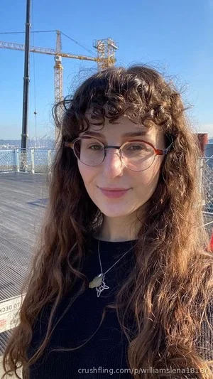 Woman with curly hair and glasses, wearing a black top, outdoors on a deck with cranes in