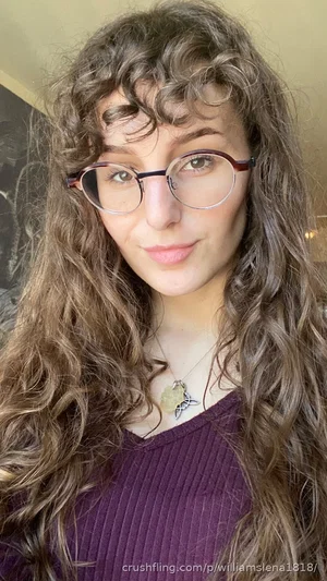 Woman with curly brown hair and glasses wearing a purple top indoors.
