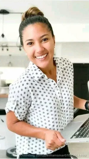 Woman in polka-dot shirt holding laptop, smiling, indoor setting.
