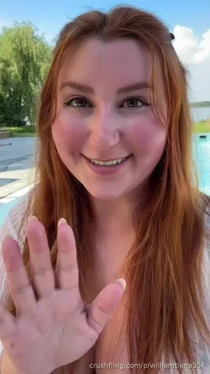 Woman with long red hair waving, wearing a sheer top, outdoors near a pool.