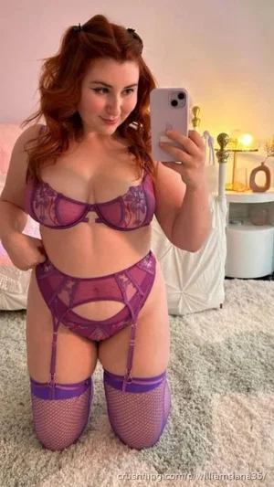 Woman with red hair in purple lingerie and fishnet stockings, kneeling on carpet in bedroom.