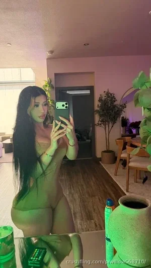 Woman with long dark hair in sheer green bodysuit taking mirror selfie in indoor living space.