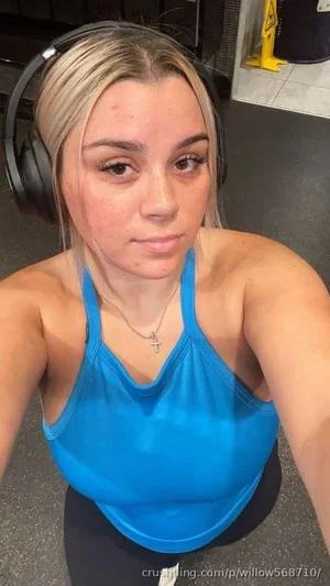 Woman with blonde hair wearing blue tank top and headphones, taking selfie in gym.