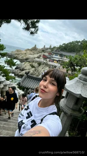 Woman with short dark hair in white graphic tee, taking selfie on stone steps near coastal temple.
