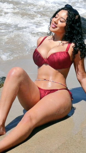 Woman in red bikini sitting on beach near waves, curly hair, smiling.