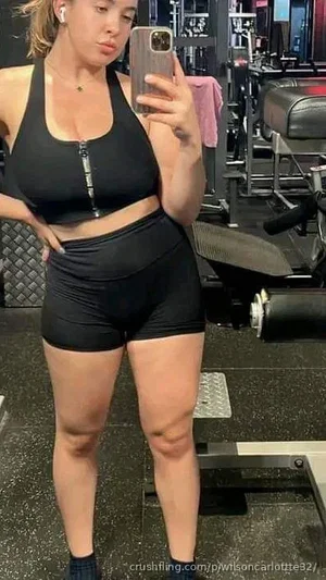 Woman in black workout top and shorts taking mirror selfie in gym.