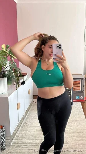 Woman in green sports bra and black leggings taking mirror selfie in bedroom.