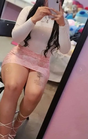 Woman with long dark hair in pink skirt and white top taking mirror selfie.