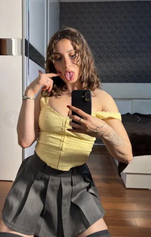Curvy woman with curly hair in yellow top taking bedroom selfie.