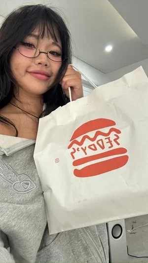 Woman with glasses holding a white bag with a red burger logo, wearing a gray hoodie indoors.
