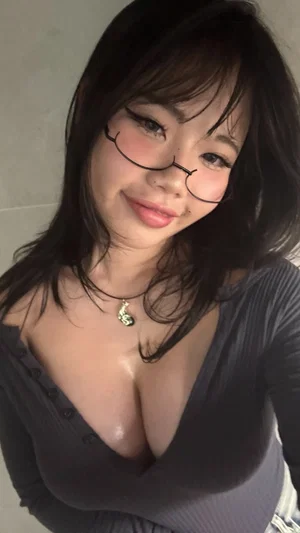 Woman with dark hair and glasses wears a low-cut gray top indoors.