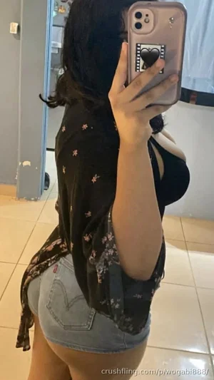 Woman in floral shirt and denim shorts taking mirror selfie indoors.