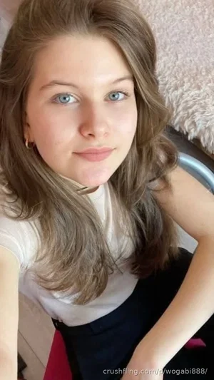 Woman with long brown hair and blue eyes wearing white top and dark skirt, sitting indoors.