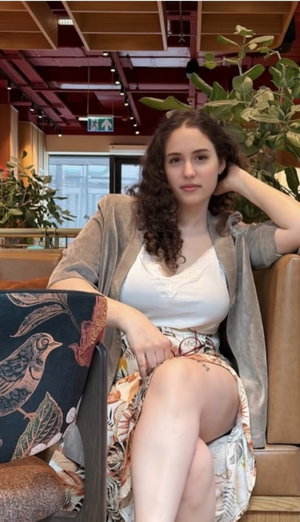 Woman with curly hair in white top and floral skirt sitting on couch indoors.