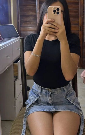 Woman with long dark hair wearing black top and denim skirt indoors.