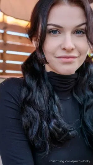 Woman with long dark hair wearing black turtleneck, outdoor setting with wooden slats.