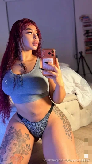 Woman with red hair and tattoos wears a blue crop top and patterned underwear, posing in a bedroom.