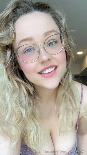 Blonde woman with glasses in a purple top, smiling in a close-up selfie indoors.