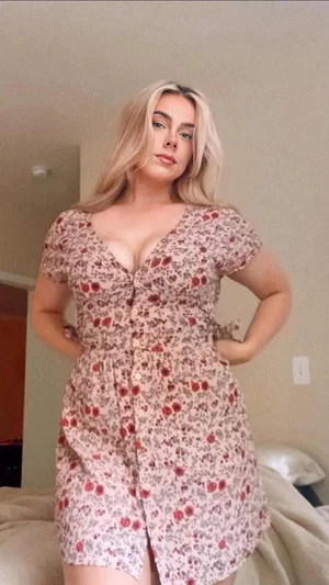 Woman with blonde hair in floral dress posing in bedroom.