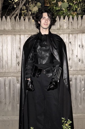 Man in black superhero costume with cape, standing outdoors by wooden fence.