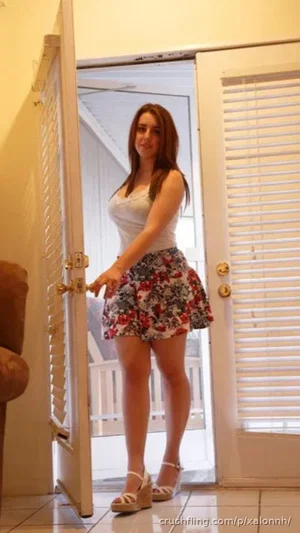 Woman in white tank top and floral skirt standing in doorway, indoor setting.