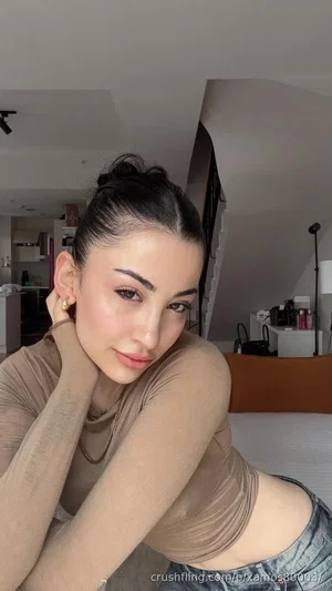 Woman with dark hair in bun, wearing beige top and jeans, posing indoors.