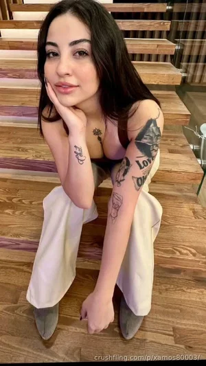 Woman with tattoos sitting on wooden steps, wearing black top and white pants.