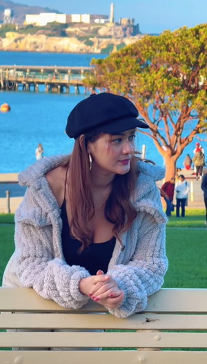 Woman in black cap and fuzzy jacket on bench by water.