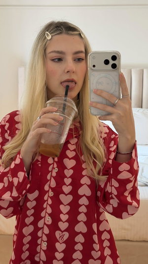 Blonde woman in red heart pajamas taking mirror selfie in bedroom.