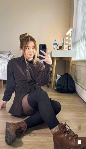 Woman in dark shirt and black tights sits on floor, taking mirror selfie in bedroom.