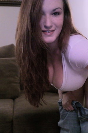 Woman with long brown hair in a tight white top, pulling down jeans, sitting on a couch indoors.