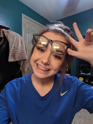 Woman with glasses and braces smiles in a blue shirt indoors.