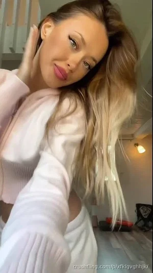 Woman with long blonde hair in pink ribbed top, posing indoors with hand on face.