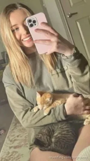 Blonde woman in olive green sweatshirt holding two kittens, taking mirror selfie indoors.